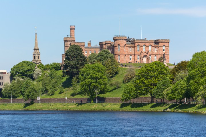 A historic red-brick castle on a green hill with a river in the foreground and a tall spire to the left.