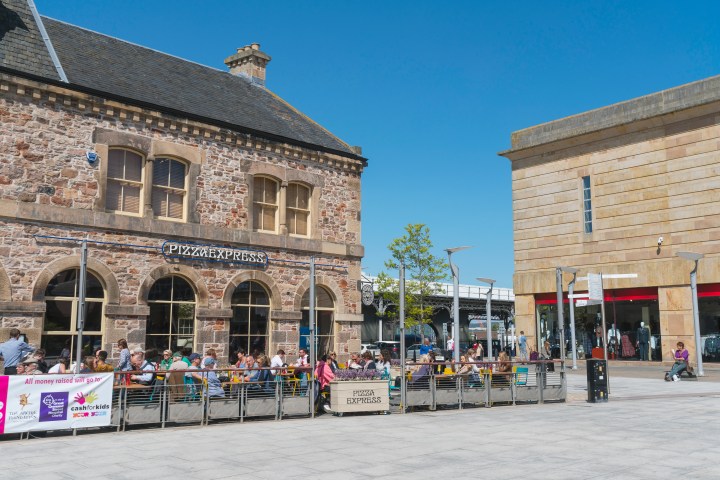 People dining outdoors at a PizzaExpress near a shopping area under a clear blue sky.