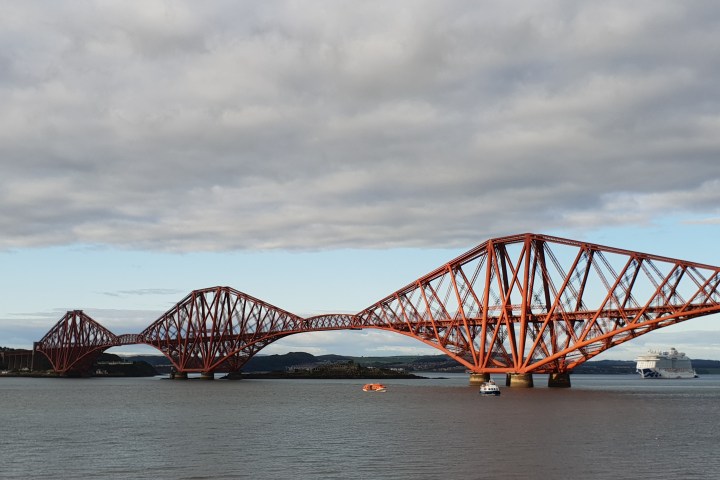 Red steel bridge over a body of water with boats and cloudy sky in the background.