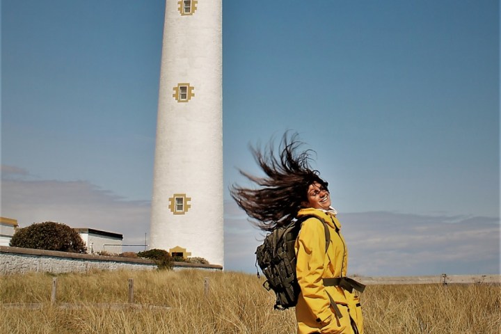 Person in yellow coat with windblown hair stands near a lighthouse on a clear day.