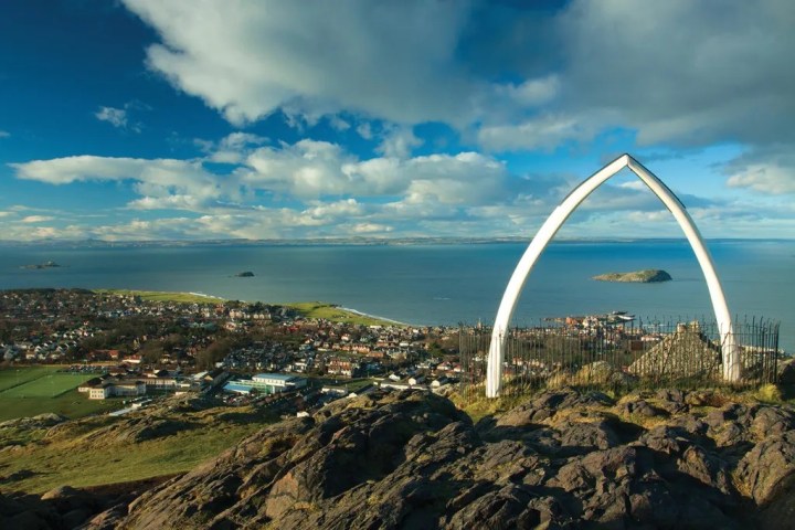 Coastal town view from hilltop with arch structure and sea in background under cloudy sky.