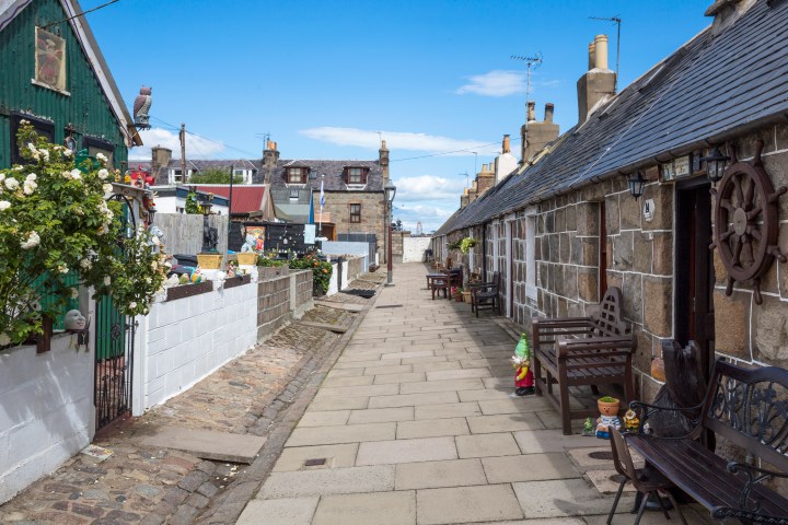 Narrow cobblestone street with quaint stone cottages and colorful garden decor.