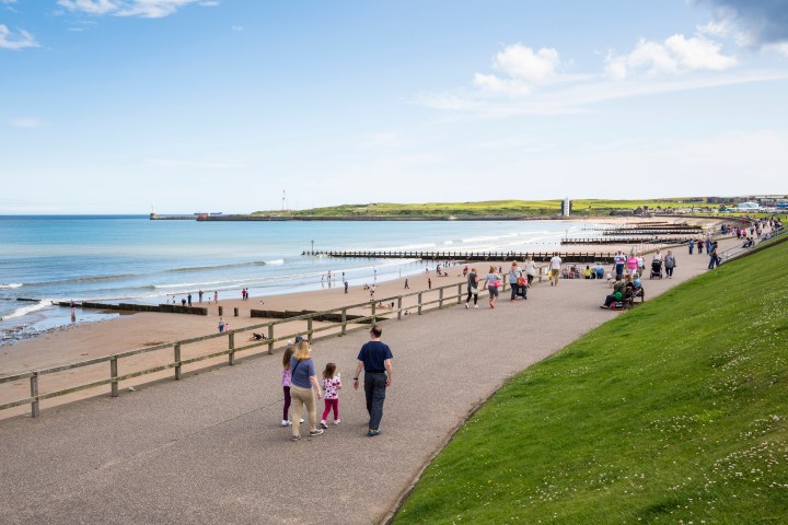 People walking on a promenade near a busy beach with sea and grassy hills in the background.