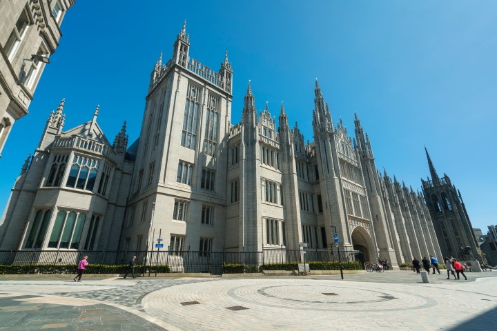 Gothic-style stone building with tall spires and people walking nearby under a clear blue sky.