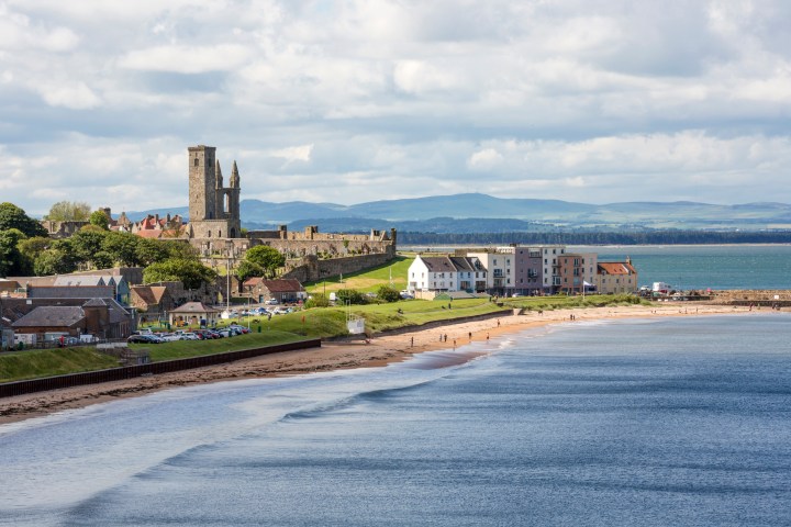 Coastal townscape with a beach, historic ruins, and buildings beside a calm sea under a cloudy sky.