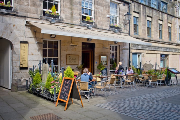 Outdoor café with tables, chairs, and people seated on a cobblestone street beside a historic building.