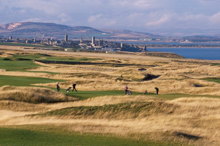 Golfers on a grassy course with a coastal town and hills in the background.