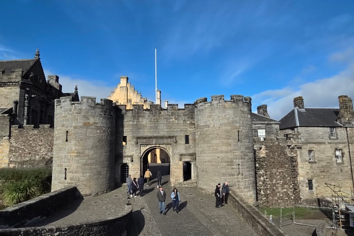 Stone castle entrance with round towers under a blue sky, people walking on pathway.