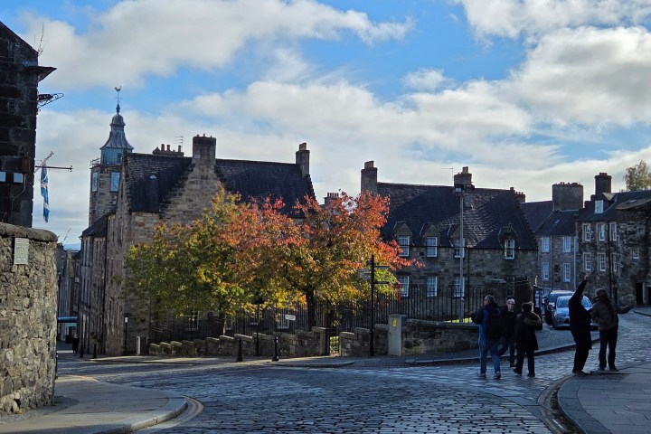Historic stone buildings and people on a cobblestone street with autumn trees.