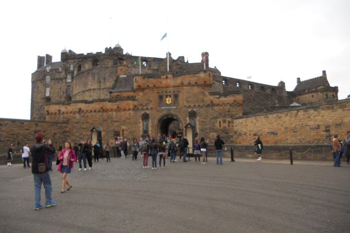 Tourists outside an ancient stone castle with flags and entry gate.