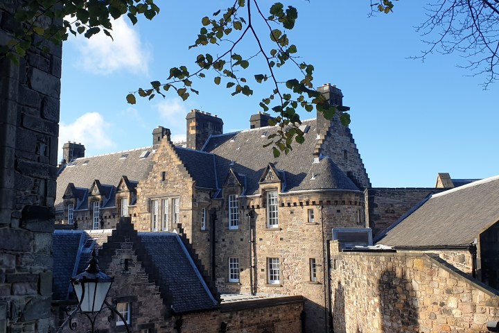 Stone building with gabled roofs under a clear blue sky, framed by branches with green leaves.