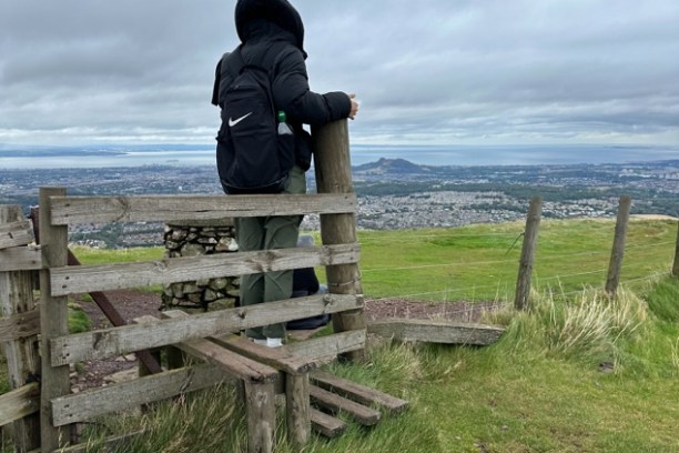 Person in black coat and backpack overlooking a city from a hillside with overcast sky.