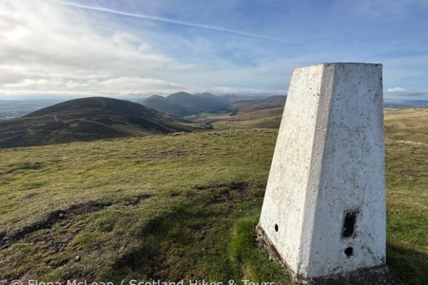 Trig point on a grassy hilltop with distant rolling hills under a cloudy sky.