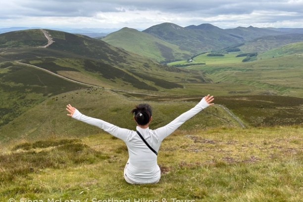 Person sitting on a grassy hill with arms wide, overlooking rolling green hills and a cloudy sky.