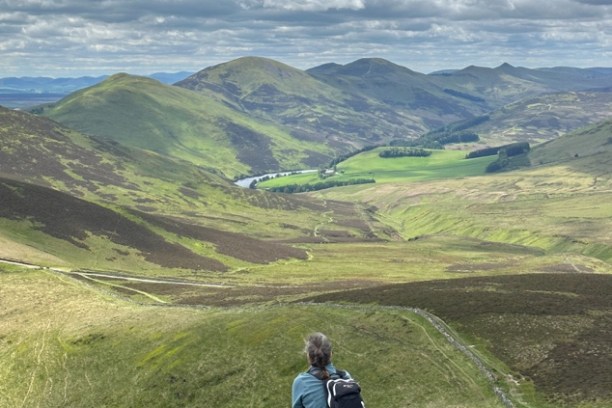 Person sitting on grassy hill, overlooking rolling green landscape and distant hills under cloudy sky.