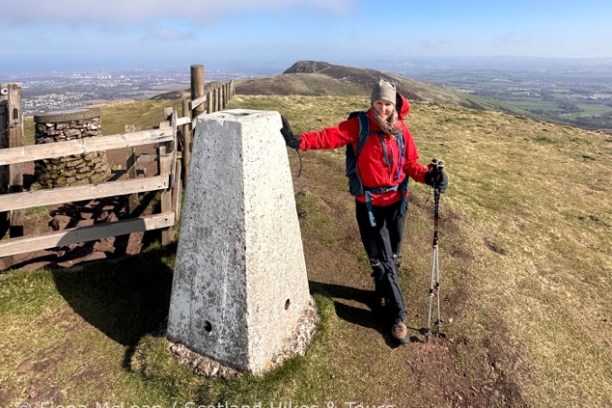Person in hiking gear stands by a stone marker on a grassy hill with a scenic view.