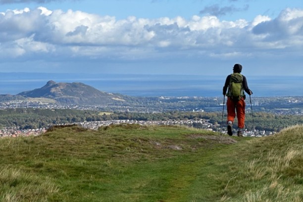 Person hiking on a hill with walking sticks, overlooking a scenic landscape and distant town.