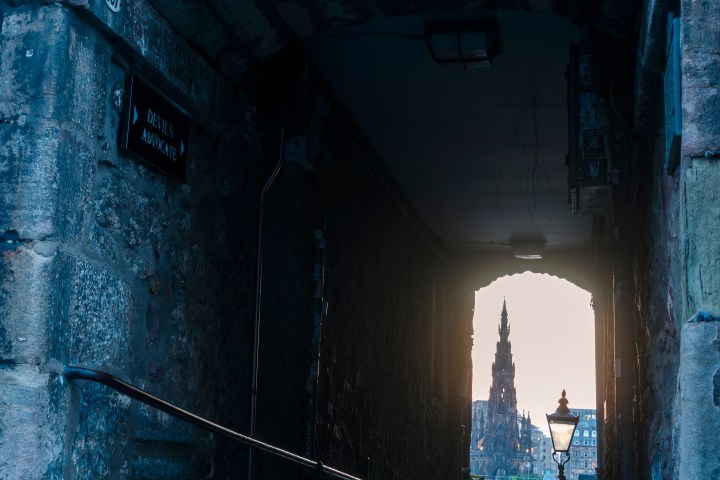 Stone archway labeled 'Advocate's Close' overlooking distant cityscape with tower at sunset.