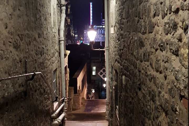 Narrow stone alley with overhead light and city view at night in distance.