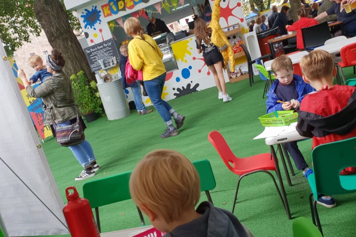 Children coloring at outdoor cafe with vivid decorations and people in background.