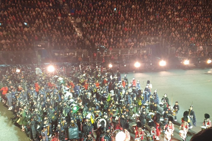 Large marching band in uniform performs in arena, audience visible.